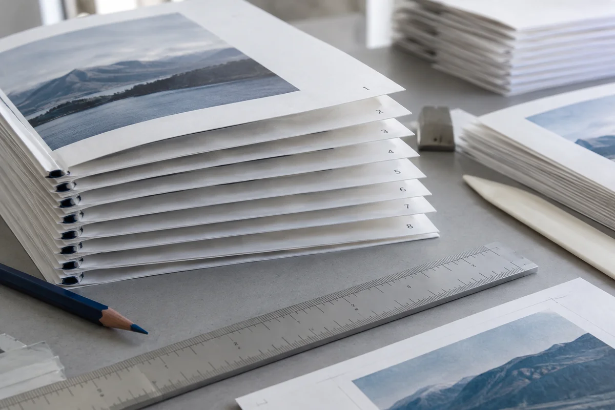 A stack of booklet signatures laid on a gray print-preparation table with tiny successive footer numbers, indigo pencil and page-layout ruler, low close view.